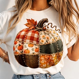 Könnte beinhalten: Ein weißes T-Shirt mit einer großen, zentralen Grafik eines Patchwork-Kürbisses. Der Kürbis besteht aus verschiedenen Stoffmustern, darunter rotes Karomuster, Blumenprints, dunkelgrünes Fischgrätenmuster und Herbstblätter, die durch sichtbare Nähte verbunden sind.