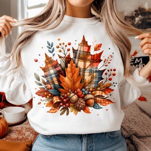 May include: A white crewneck sweatshirt featuring a central graphic of autumn foliage, including plaid maple leaves in shades of orange, red, and blue, accompanied by acorns, pinecones, and small berries, all arranged in a decorative cluster.