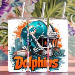 May include: White tumbler with a colorful graphic of a football helmet with a dolphin logo and the word "Dolphins" in orange. The background is a white and orange splatter design.