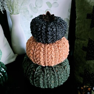 May include: A stack of three knitted pumpkins in fall colors. The top pumpkin is dark gray with a brown leaf accent. The middle pumpkin is orange, and the bottom pumpkin is green. The pumpkins are stacked on top of each other.