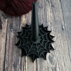 May include: A black yarn swift with a spiderweb design. The swift has a central cone and a circular base with a web pattern. A burgundy ball of yarn is partially visible in the background. The swift is designed for yarn organization.