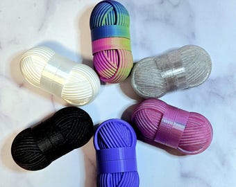 May include: Six small yarn holders in various colours, including white, black, purple, pink, and a rainbow pattern. The holders are shaped like balls of yarn and have a textured surface.