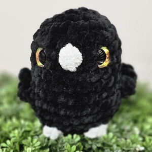 May include: A handmade black plush bird with large, iridescent eyes and a white beak. The bird is made of a soft, textured material and sits on a bed of green foliage. A charming, whimsical stuffed animal.