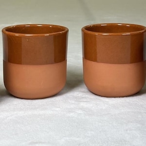 Puede incluir: Cuatro tazas de cerámica con un diseño de dos tonos. La mitad superior de cada taza es de color marrón oscuro, mientras que la mitad inferior es de color terracota más claro. Las tazas tienen una forma redondeada y un acabado liso.
