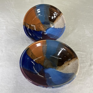 Può includere: Due ciotole in ceramica con un design astratto e colorato. Le ciotole presentano un mix di colori, tra cui sfumature di blu, marrone e arancione. Le ciotole sono rotonde con una finitura lucida e il design è applicato a segmenti.