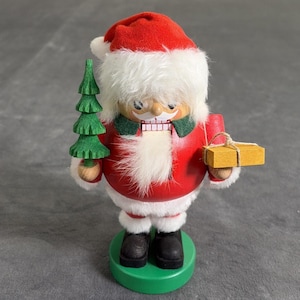 May include: A wooden nutcracker figurine depicting a figure in a red jacket and hat with white faux fur trim. The figure has a white beard, holds a small green wooden Christmas tree in one hand, and a rectangular yellow gift box in the other.