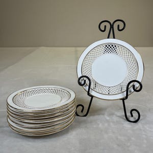 Puede incluir: Un conjunto de platos de porcelana blanca con borde dorado y diseño de celosía. Un plato se exhibe en un soporte de metal negro. Los platos son circulares y tienen un acabado liso y brillante. Adecuado para postres.