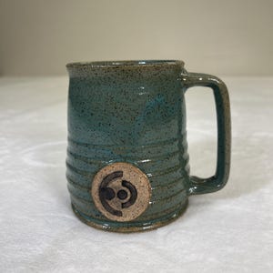 Może przedstawiać: Turkusowy ceramiczny kubek z uchwytem i okrągłym emblematem. Kubek ma nakrapianą fakturę i poziome żłobienia. Emblemat jest w jasnobrązowym kolorze i ma stylizowany wzór.