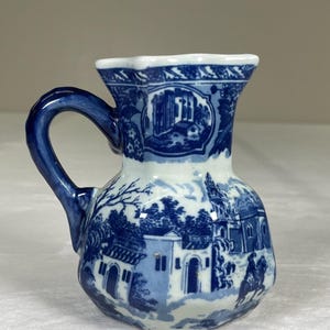 Könnte beinhalten: Kleine blau-weiße Keramikkrug mit Henkel. Der Krug zeigt eine detaillierte Landschaftsszene mit Gebäuden und Bäumen. Der Rand und der Henkel sind dunkler blau, und das Gesamtdesign erinnert an antikes Porzellan.