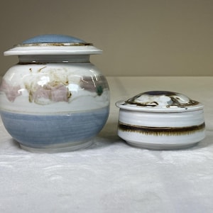 Puede incluir: Dos tarros de cerámica con tapa. El tarro más grande tiene una base y tapa azul claro, con detalles blancos y florales. El tarro más pequeño es blanco con una banda marrón y una tapa marrón oscuro.