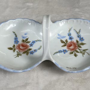 May include: A white, divided serving dish with a handle, featuring a floral design. The dish has a light blue scalloped edge and is decorated with pink roses and blue flowers. The handle is white, and the dish is likely made of ceramic.