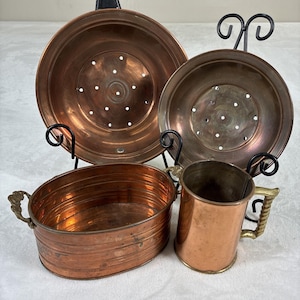 May include: A collection of vintage copper kitchenware, including two colanders, an oval basin with ornate handles, and a mug with a brass handle. The items are displayed on black metal stands, showcasing their aged patina and rich copper tones.