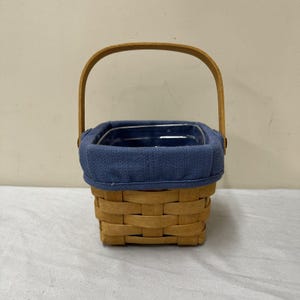 Puede incluir: Una pequeña cesta cuadrada tejida con un asa de madera. La cesta tiene un forro de tela azul y una inserción de plástico transparente. La cesta está hecha de material tejido marrón claro.