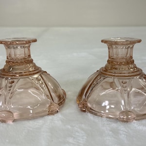 Könnte beinhalten: Zwei passende rosafarbene Glas-Kerzenhalter. Jeder hat einen runden Sockel mit einem dekorativen Muster und einen schlanken, geriffelten Hals. Die Kerzenhalter bestehen aus durchscheinendem rosa Glas.