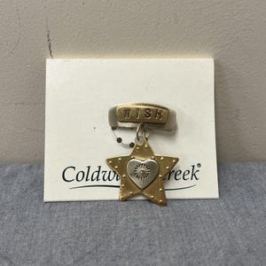 May include: A gold and silver-toned metal brooch with a star and heart design. The brooch has the word "WISH" on a silver bar. The star has a heart in the center with a floral design. The brooch is attached to a white card with the words "Coldwater Creek".