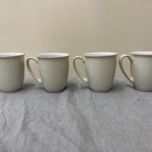 May include: Four off-white ceramic mugs with curved handles are arranged in a row. The mugs have a simple, classic design with a slightly flared shape and a smooth, matte finish. The interior of the mugs is white.