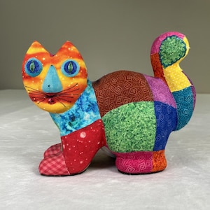 May include: A colorful patchwork cat figurine. The cat features a vibrant mix of fabrics in orange, blue, red, green, and brown. The cat's face has large blue eyes and a blue nose. The tail is upright, and the cat is posed as if it is walking.