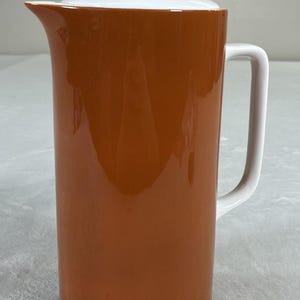 Puede incluir: Una jarra de cerámica naranja, cilíndrica y alta, con asa, boquilla y tapa blancas. La jarra tiene un acabado brillante y un diseño sencillo y moderno. Ideal para servir bebidas.