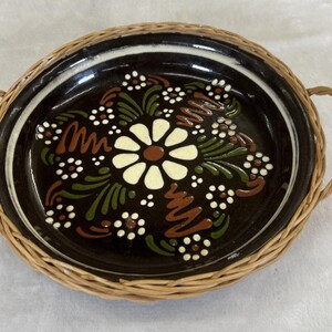 May include: Round, dark brown ceramic tray with a woven wicker border and handles. The tray features a floral design in white, green, and brown. The tray is approximately 10 inches in diameter.
