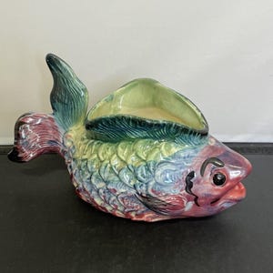 May include: A ceramic fish-shaped planter in a vibrant array of colours. The planter features a detailed scale pattern and a wide opening on its back for plants. The fins and tail are intricately designed. The fish is painted with blues, greens, and reds.