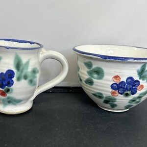 May include: A white ceramic mug and bowl with a blue and green floral design. The design features blue berries and green leaves.