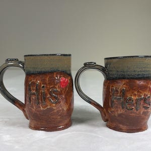 May include: Two handmade ceramic mugs. One mug reads "His" and the other reads "Hers" in a textured, brown glaze. Each mug has a dark handle and a red heart accent. The top rim of each mug is a darker color.