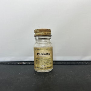 May include: A small, clear glass bottle with a rusty metal lid. The label reads "Phenolax" and lists the ingredients as Phenolphthalein, Ginger and aromatics. The bottle is labelled "10 Wafers" and "No. 6035".