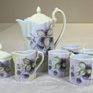 Puede incluir: Juego de té de porcelana vintage con tetera, azucarero, jarrita de leche y cuatro tazas. El juego está decorado con moras moradas y hojas verdes sobre un fondo lavanda pálido. La tetera mide aproximadamente 20 cm de alto.