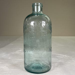 May include: A vintage, clear glass bottle with embossed text. The bottle is a light aqua color and features the text "Sand Lake Springs" and "West Sand Lake, New York". The bottle is approximately 9 inches tall.