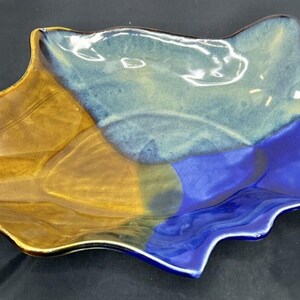 May include: A ceramic leaf-shaped dish with a glossy finish. The dish is divided into sections of brown, blue, and teal. The edges are wavy, and the dish sits on a dark surface.