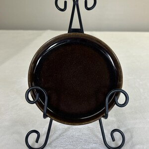 Peut inclure: Une petite assiette ronde en céramique brun foncé présentée sur un chevalet en métal noir. L'assiette a une finition brillante et un bord légèrement surélevé. Le chevalet présente des volutes décoratives en haut et en bas.