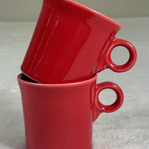 May include: Two stacked, glossy red ceramic mugs. Each mug has a rounded handle and a slightly flared rim. The mugs are a vibrant, solid red color, and the handles are circular.