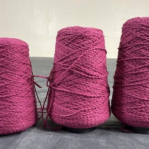 May include: Three conical spools of magenta yarn arranged in a row. Each spool is densely wound with a textured, thick-ply magenta thread, showing a consistent deep pink color throughout the entire set of three individual yarn cones.