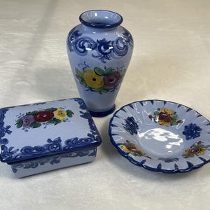 May include: A set of three blue and white ceramic pieces with floral designs. The set includes a vase, a small bowl, and a square box with a lid.