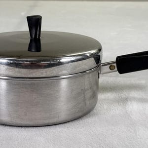 May include: A stainless steel saucepan with a lid and a black handle. The lid has a black knob. The pan is sitting on a white surface. The handle has a metal loop at the end.