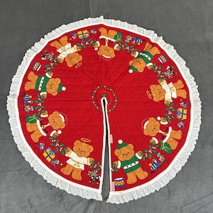 May include: A circular red quilted Christmas tree skirt featuring a repeating pattern of brown teddy bears wearing green sweaters or white angel robes, interspersed with colorful gift boxes, toy trains, and green holly leaves with a white lace trim.