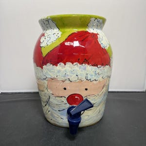 Könnte beinhalten: Ein Keramik-Getränke-Spender mit einem Weihnachtsmann-Design. Der Spender ist grün, rot, weiß und blau. Er hat einen blauen Hahn.