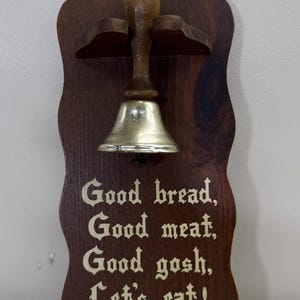 Puede incluir: Una campana de madera para colgar en la pared, con un acabado marrón oscuro. La campana tiene un badajo plateado y un mango de madera. El letrero dice: "Good bread, Good meat, Good gosh, Let's eat!"