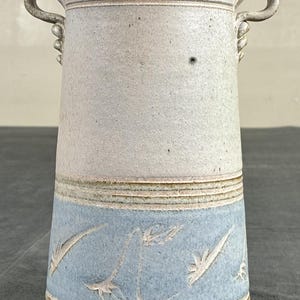 May include: A tall, cylindrical ceramic vase with two small, curved handles at the top. The upper portion is a speckled off-white, while the lower section features a light blue band decorated with incised, stylized feather or leaf patterns.