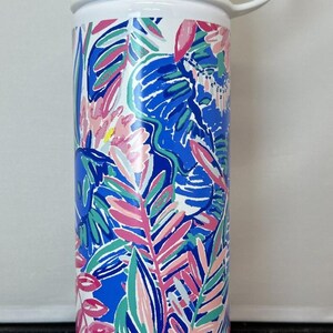 May include: White water bottle with a stainless steel lid and handle. The bottle features a colourful floral design in blue, pink, and green. The bottle is approximately 25 cm tall.