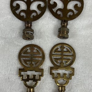 May include: Four brass lamp finials. Two have a circular design with an open, swirling pattern. The other two have a rectangular base with a Chinese character design. All have a threaded base for attachment.