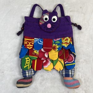 Può includere: Una borsa tote a forma di gatto fatta a mano. La borsa presenta un corpo, un viso e manici viola. La parte inferiore è decorata con scaglie di tessuto colorate e fantasia. La borsa ha due piedi in tessuto patchwork. Un accessorio unico e giocoso.