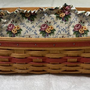 May include: Rectangular woven basket with a floral fabric lining. The basket is made of light brown and red woven strips. The top edge is trimmed with a red band. The interior lining features a floral pattern with pink and yellow roses.