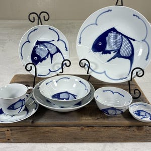 Puede incluir: Un conjunto de vajilla de cerámica blanca con un diseño de pez azul. El juego incluye platos, cuencos, una taza y un platillo, y un plato pequeño. Los peces son estilizados y el conjunto se exhibe sobre una superficie de madera.