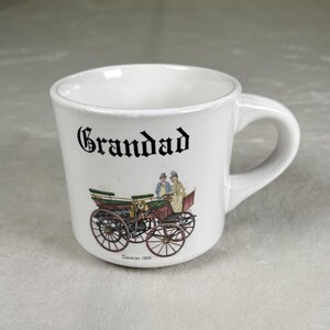 Può includere: Tazza in ceramica bianca con la scritta "Grandad" in nero. La tazza presenta un'illustrazione colorata di un'auto d'epoca con due figure. L'auto è rossa, verde e marrone, con il testo "Daimler 1886" sotto.