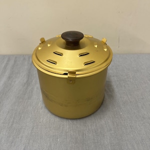 Może przedstawiać: Złoty metalowy garnek do gotowania z pokrywką. Pokrywka ma ciemnobrązowy drewniany uchwyt i kilka szczelin wentylacyjnych. Garnek ma cylindryczny kształt z uchwytem z boku. Garnek stoi na szarej powierzchni.