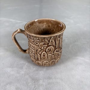 May include: A brown ceramic coffee mug with a textured surface. The mug features embossed lettering spelling out "Kaffe", "Coffee", and other words. The handle is a branch-like design.