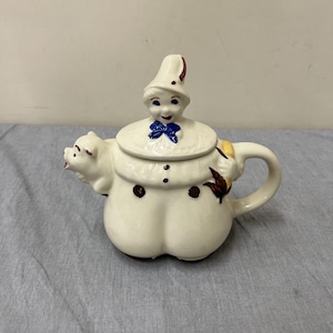 Puede incluir: Tetera de cerámica de color crema, caprichosa, con un pico en forma de cabeza de cerdo y una tapa con una figura sonriente con sombrero y pajarita azul. La tetera tiene un cuerpo redondeado con detalles de botones marrones y un asa curvada.