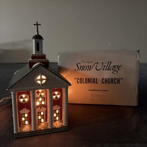 May include: A ceramic Colonial Church building from The Original Snow Village with a red brick facade, white columns, and a green roof. The structure is illuminated from within, featuring a cross on the steeple and its original white cardboard box.