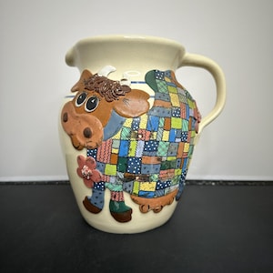 Op de afbeelding: Crèmekleurige keramische kan met een grillig koeienontwerp. De koe heeft een patchworkpatroon in verschillende kleuren, bruine oren en een bloemaccent. De kan heeft een handvat en een tuit.